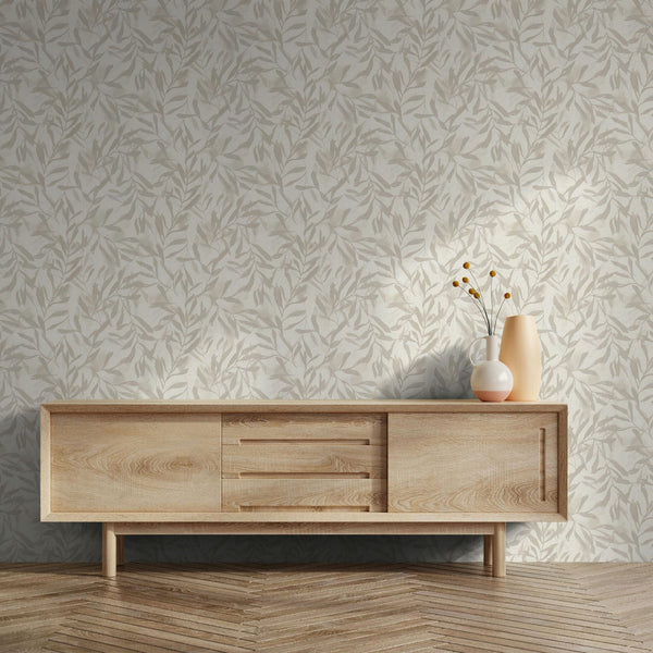 Marburg Leyla 82441 Beige wallpaper with an all-over leafy pattern behind a light wood sideboard topped with two vases and delicate stems, on a herringbone wood floor.