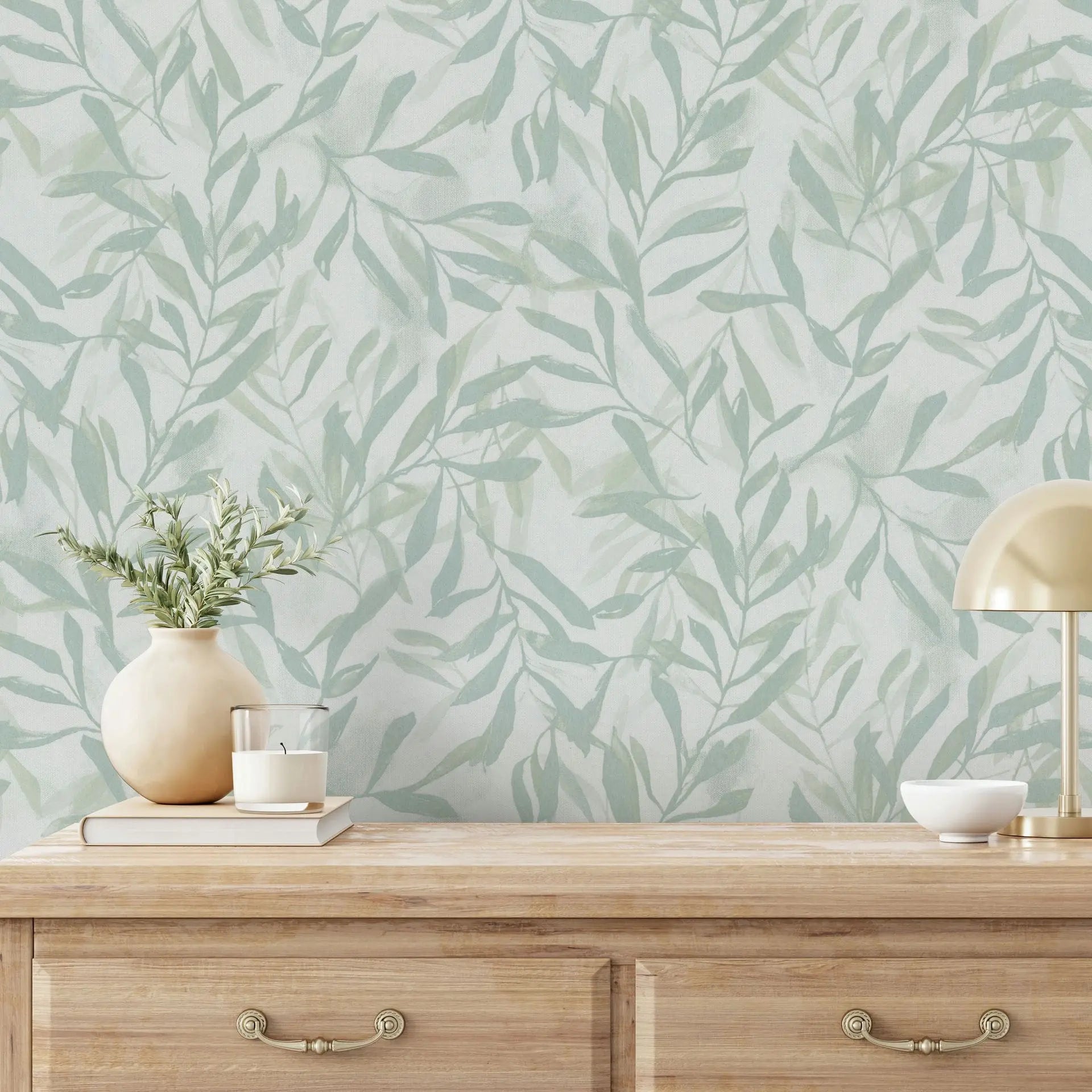 Marburg Leyla 82442 Light green leafy wallpaper behind a wooden console table with a small brass desk lamp, a vase with foliage, stacked books with a candle, and a small bowl.