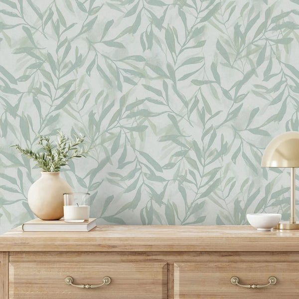 Marburg Leyla 82442 Light green leafy wallpaper behind a wooden console table with a small brass desk lamp, a vase with foliage, stacked books with a candle, and a small bowl.