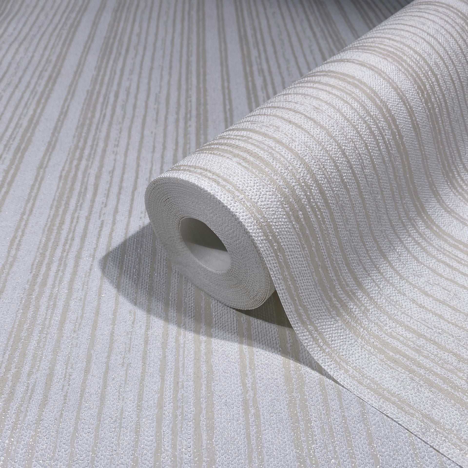 Marburg Leyla 82443 Close-up of a wallpaper roll with beige and white irregular vertical stripes and a textured surface, resting on a matching unrolled sheet.