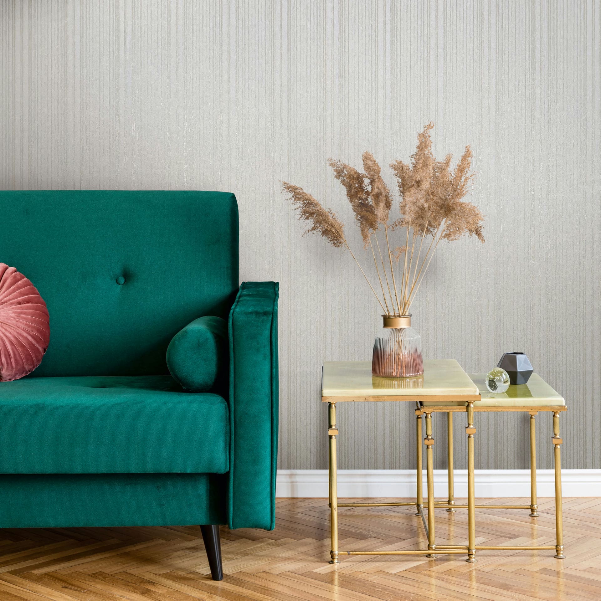 Marburg Leyla 82443 Light beige wallpaper with fine vertical striations behind a green sofa and gold nesting tables holding a vase of pampas grass.