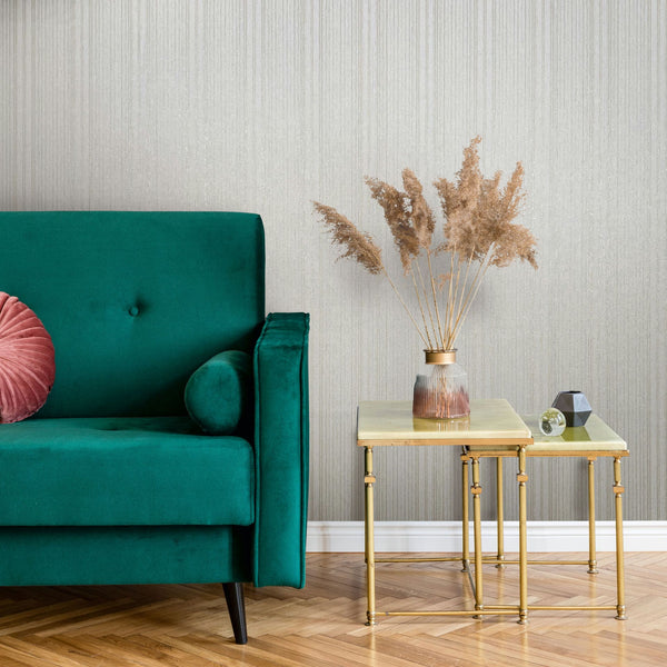 Marburg Leyla 82443 Light beige wallpaper with fine vertical striations behind a green sofa and gold nesting tables holding a vase of pampas grass.