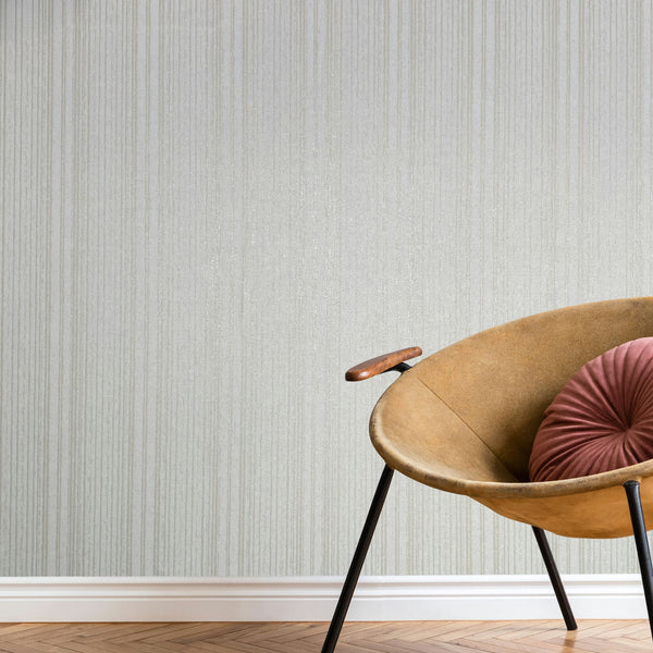 Marburg Leyla 82443 Light neutral vertical stripe wallpaper behind a tan curved lounge chair with a round pleated cushion, white baseboard, and herringbone wood floor.