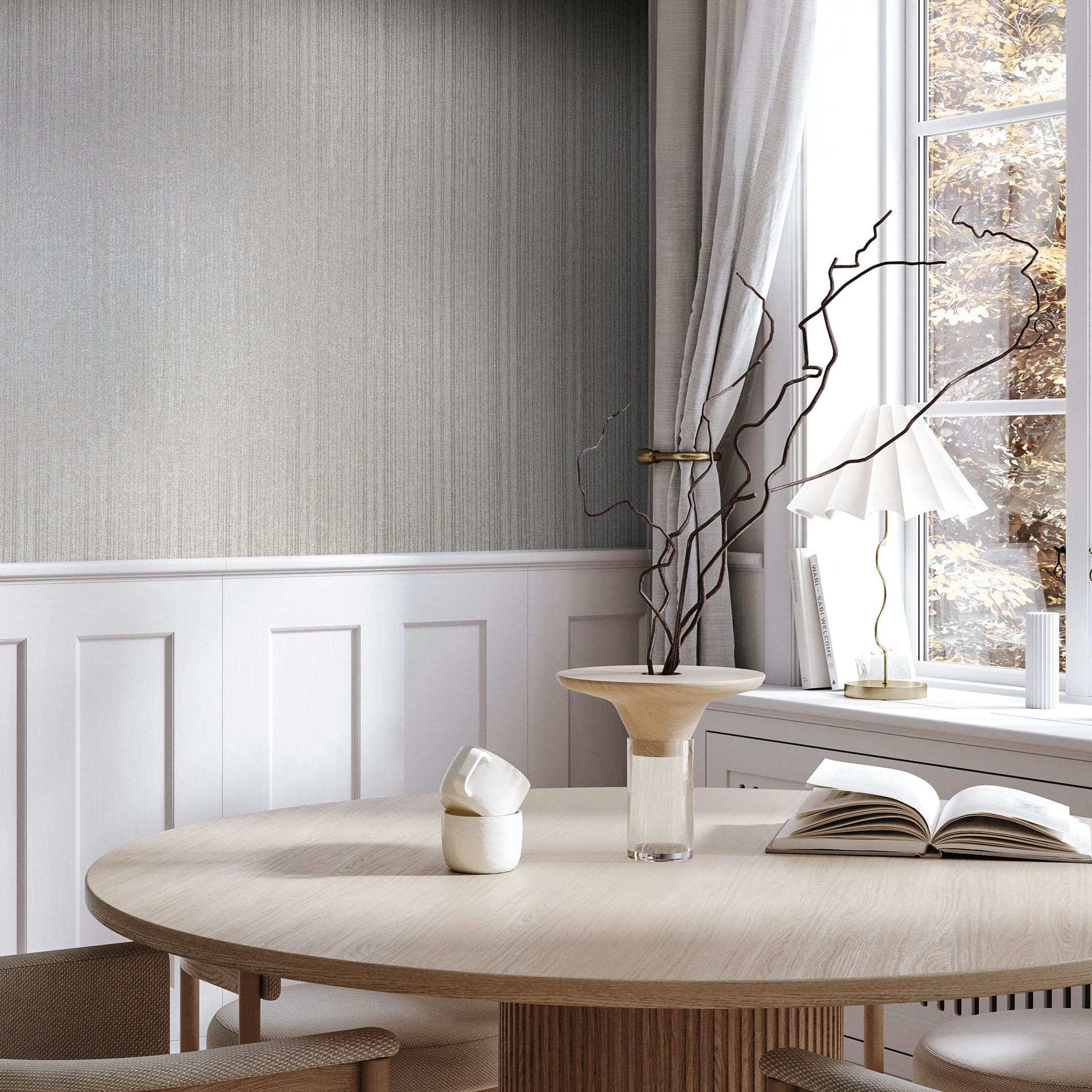 Marburg Leyla 82444 Gray vertical strié wallpaper above white wainscoting beside a window with curtains, shown with a round wood dining table, a vase with branches, an open book, and a small table lamp.
