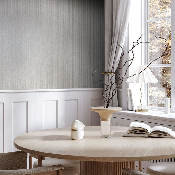 Marburg Leyla 82444 Gray vertical strié wallpaper above white wainscoting beside a window with curtains, shown with a round wood dining table, a vase with branches, an open book, and a small table lamp.