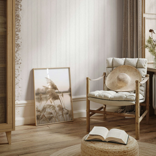 Marburg Leyla 82445 Light vertical stripe wallpaper behind a cushioned wooden chair with a straw hat, a framed photo leaning on the wall, curtains by a window, and a woven pouf with an open book.