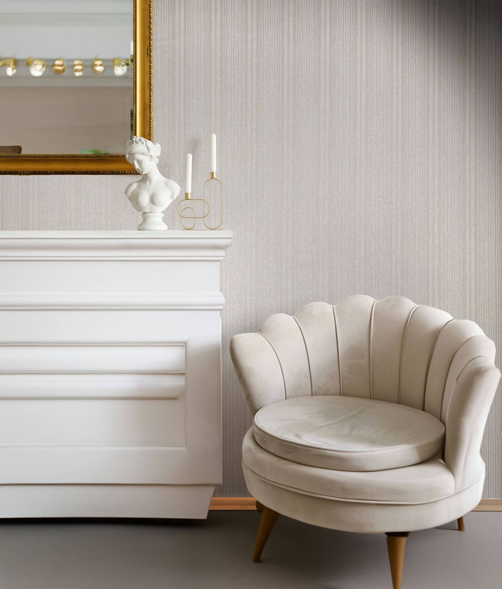 Marburg Leyla 82446 Beige vertical strie wallpaper behind a white mantel topped with a bust sculpture and two white candles in a gold holder, with a gold-framed mirror above and a cream scalloped armchair in front.