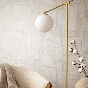 Marburg Leyla 82450 Beige abstract line-pattern wallpaper behind a brass floor lamp with a white globe shade, a textured armchair with a throw, and cotton stems.