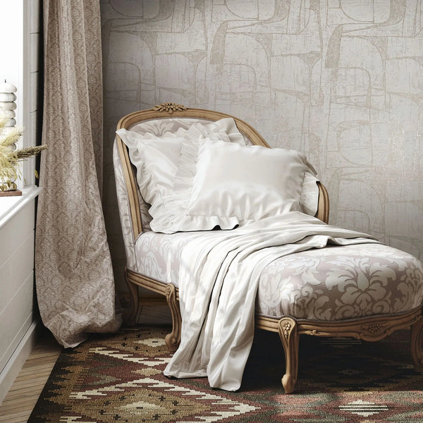 Marburg Leyla 82450 Light beige abstract geometric wallpaper behind a carved chaise with white pillows and a draped blanket beside a window with a patterned curtain and a woven rug.