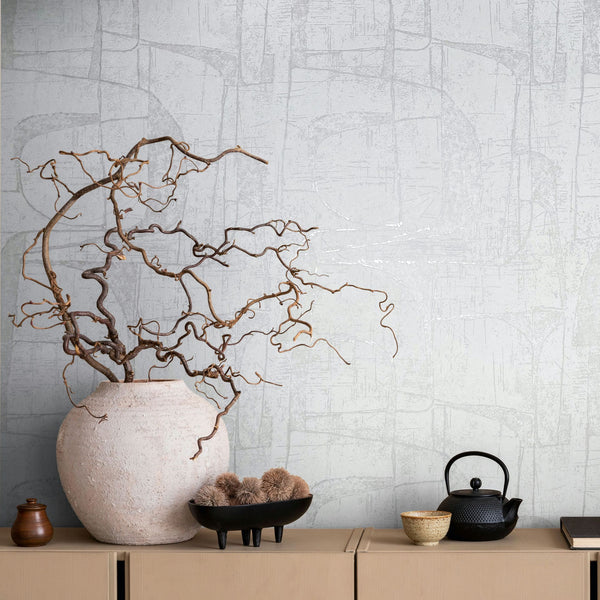 Marburg Leyla 82451 Light neutral wallpaper with a tonal abstract linear pattern behind a cabinet styled with a large textured vase of twisting branches, a small jar, a black footed bowl with dried botanicals, a ceramic cup, a black teapot, and a book.