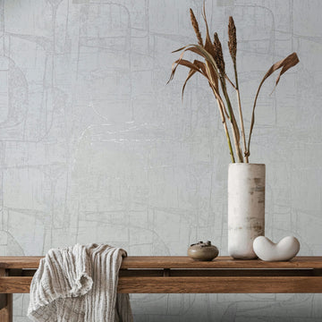 Marburg Leyla 82451 Light neutral abstract wallpaper behind a wooden bench styled with a draped throw, a tall ceramic vase holding dried stems, a small jar, and a white sculptural object.