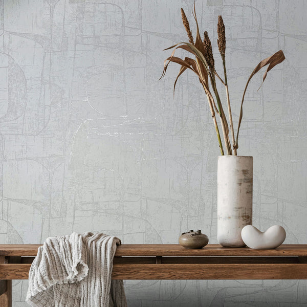 Marburg Leyla 82451 Light neutral abstract wallpaper behind a wooden bench styled with a draped throw, a tall ceramic vase holding dried stems, a small jar, and a white sculptural object.