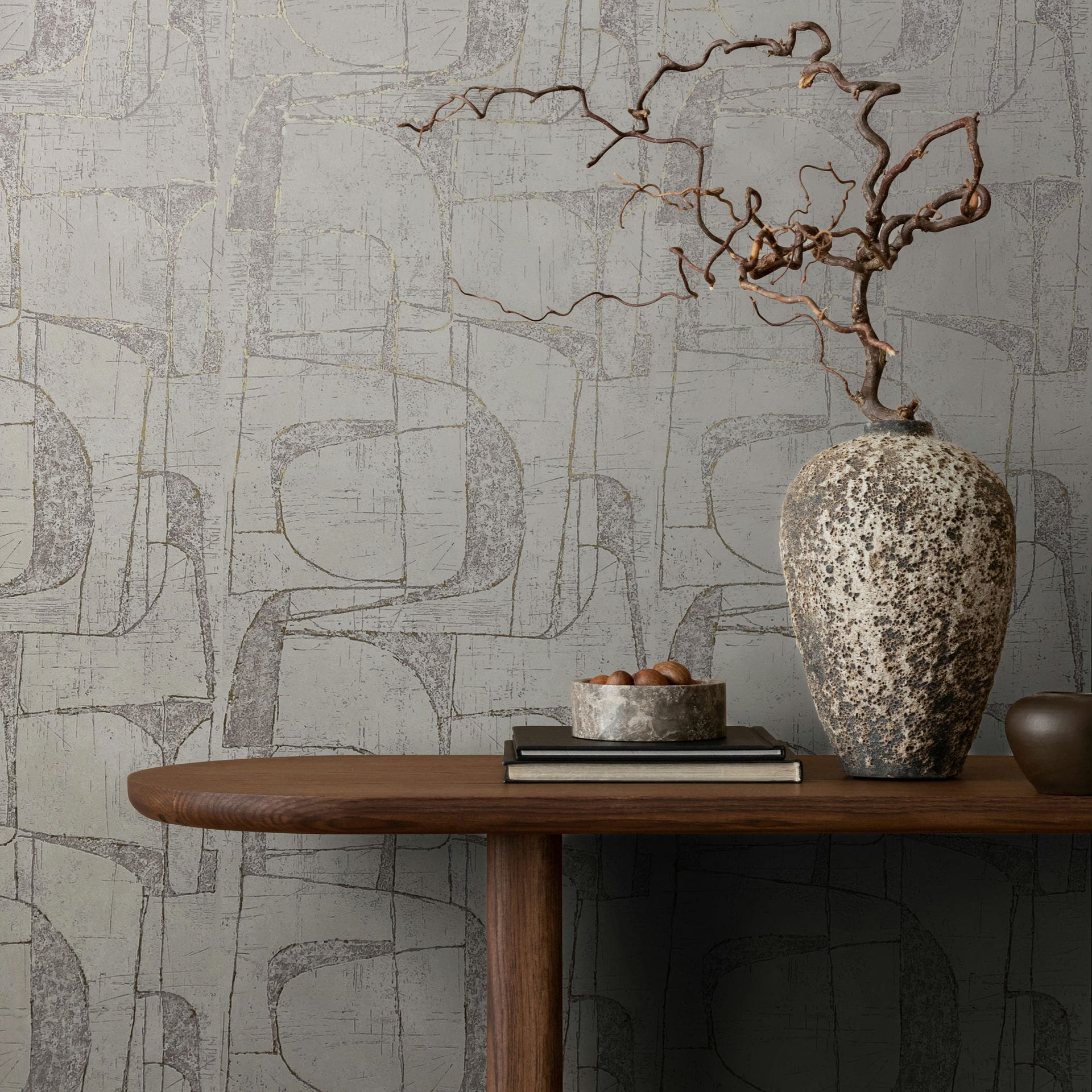 Marburg Leyla 82453 Abstract geometric wallpaper with overlapping curved shapes and a distressed texture behind a wooden console table holding a large textured vase with a dried branch, a bowl, and stacked books.