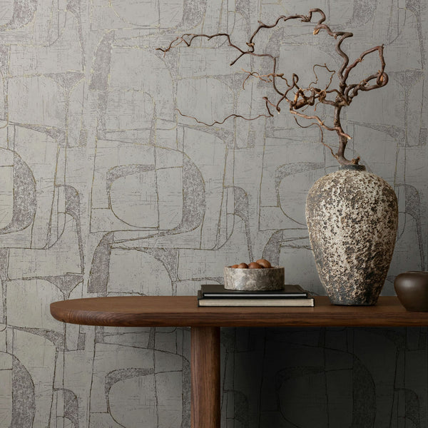 Marburg Leyla 82453 Abstract geometric wallpaper with overlapping curved shapes and a distressed texture behind a wooden console table holding a large textured vase with a dried branch, a bowl, and stacked books.