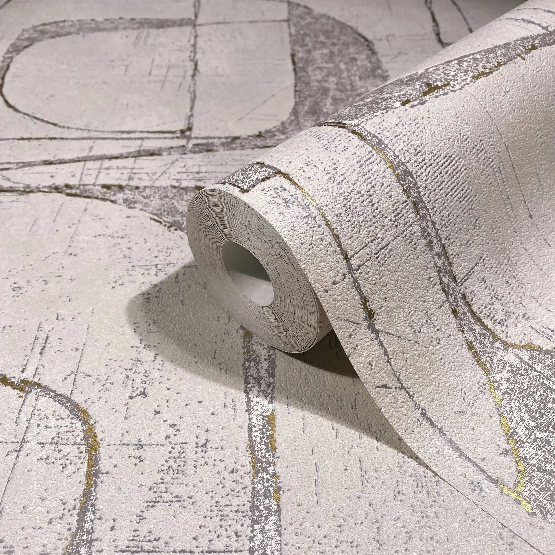 Marburg Leyla 82453 Rolled wallpaper resting on a matching unrolled sheet, showing a textured light beige surface with abstract intersecting gray lines and thin gold accents.