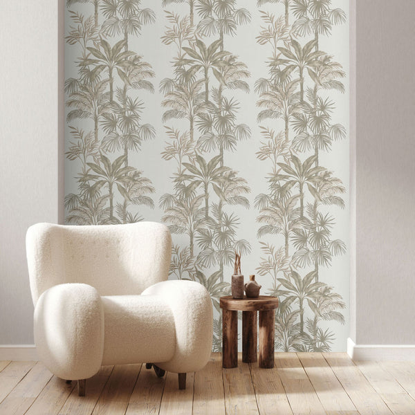 Marburg Leyla 82454 Feature wall with beige palm tree patterned wallpaper on a light background, shown behind a cream armchair and a small round wooden side table with decorative objects on wood flooring.