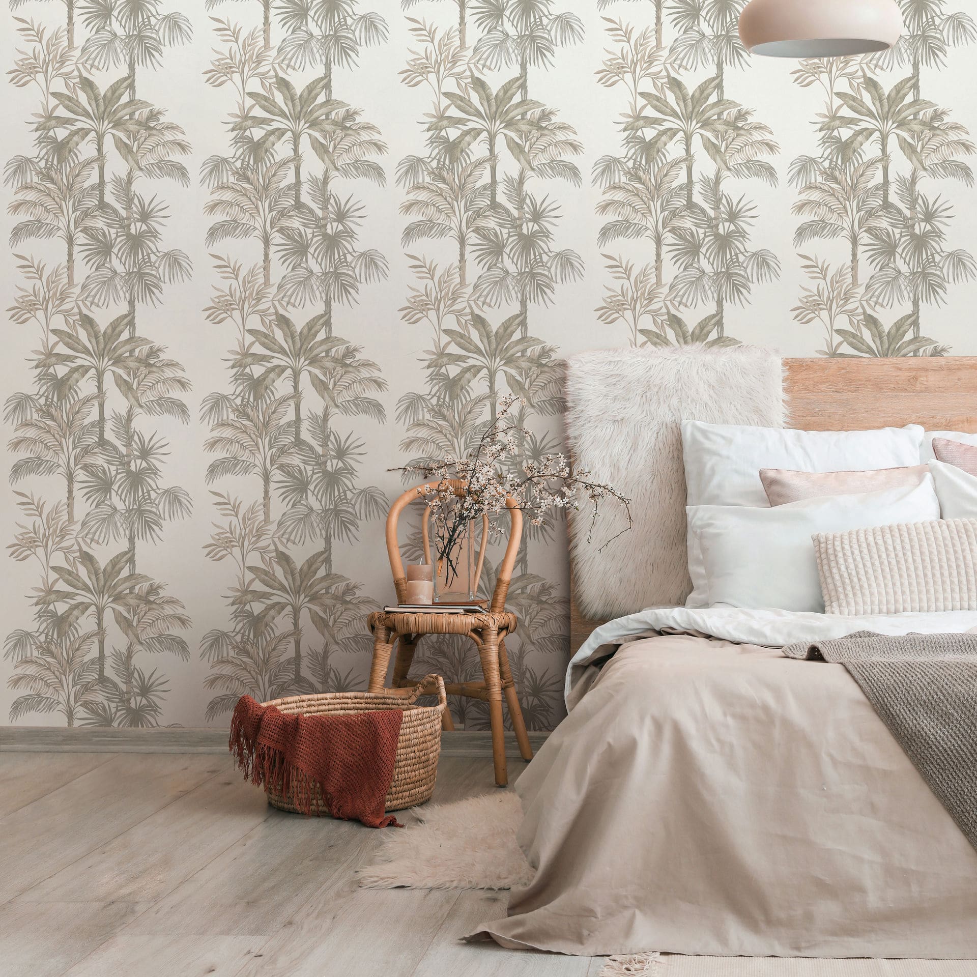 Marburg Leyla 82454 Bedroom scene with beige palm tree wallpaper behind a bed with layered pillows. A rattan chair holds a cup with dried branches beside a woven basket on a light floor, with a pendant light overhead.