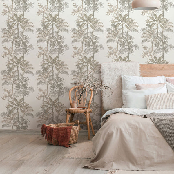 Marburg Leyla 82454 Bedroom scene with beige palm tree wallpaper behind a bed with layered pillows. A rattan chair holds a cup with dried branches beside a woven basket on a light floor, with a pendant light overhead.