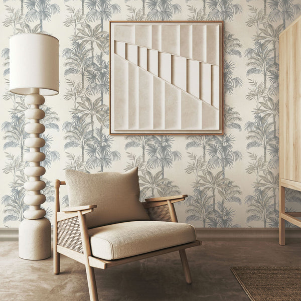 Marburg Leyla 82455 Beige wallpaper with soft blue‑gray palm trees shown in a living room setting with a lounge chair, sculptural floor lamp, framed wall art, and a wooden cabinet edge.