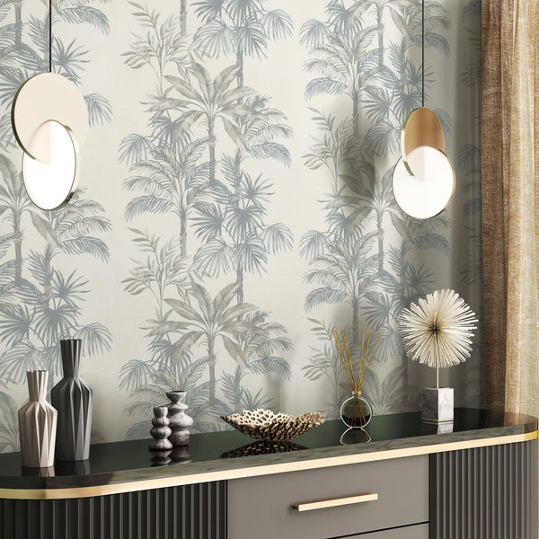 Marburg Leyla 82455 Tropical palm tree wallpaper in soft neutrals behind a sideboard with vases, a patterned bowl, a reed diffuser, a spiky sculpture, and two round pendant lights, with a curtain at right.