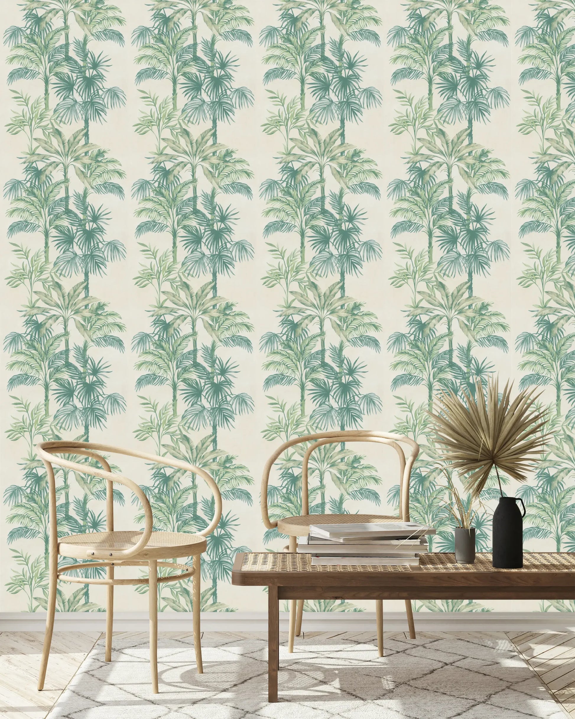 Marburg Leyla 82456 Wallpaper with repeating green palm trees on a light beige background, shown in a living room setup with two light wood bentwood chairs, a woven-top low table, stacked books, a small vase with dried palm leaves, a black pitcher, and a rug.