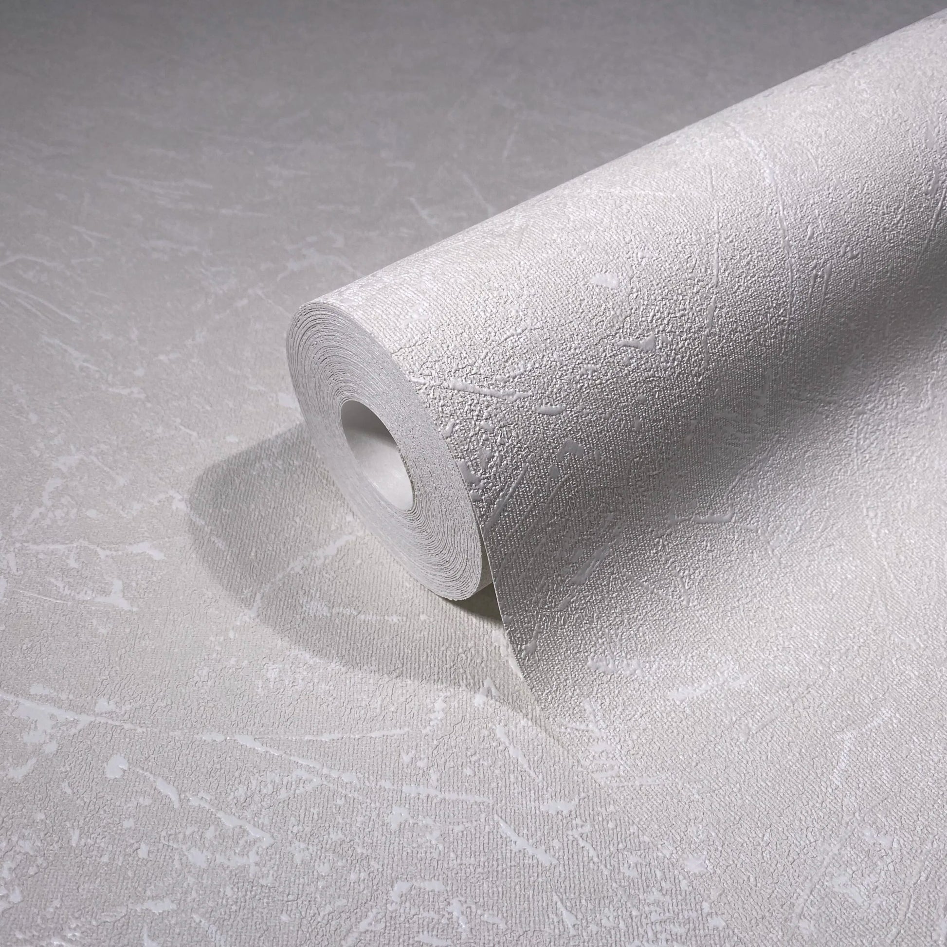 Marburg Loft Superior 34151 A wallpaper roll resting on matching unrolled wallpaper, showing a light neutral, textured surface with irregular abstract lines.