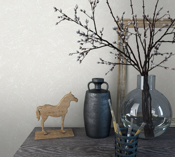 Marburg Loft Superior 34151 Light beige textured wallpaper with fine plaster-like markings shown behind a vignette featuring a clear round glass vase of branches, a dark jug, a wooden horse figurine, a leaning frame, and a cup holding paintbrushes on a grey wood table.