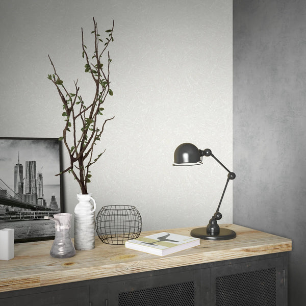 Marburg Loft Superior 34151 Light neutral textured wallpaper with fine crosshatch lines shown behind a console holding a black task lamp, a white vase with a branch, a wire basket, a glass vessel, two small white rectangular decor pieces, and a framed black‑and‑white city photograph.