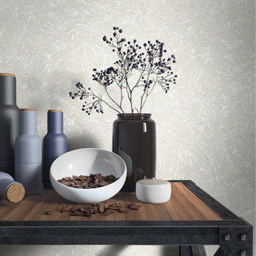 Marburg Loft Superior 34152 Light beige‑gray textured wallpaper with subtle abstract scratch lines behind a wood‑and‑metal table styled with matte bottles, a dark vase holding berry branches, a white bowl filled with beans, and a small lidded jar.