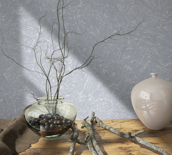 Marburg Loft Superior 34153 Gray textured wallpaper with an abstract scraped look behind a wood tabletop styled with a clear glass vase holding branches, loose twigs, and a glossy ceramic jar in sunlight.