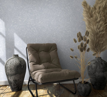 Marburg Loft Superior 34153 Gray wallpaper with an abstract scratch texture shown behind a cushioned lounge chair, two dark vases, a small pot with dried grasses, and wood plank flooring.