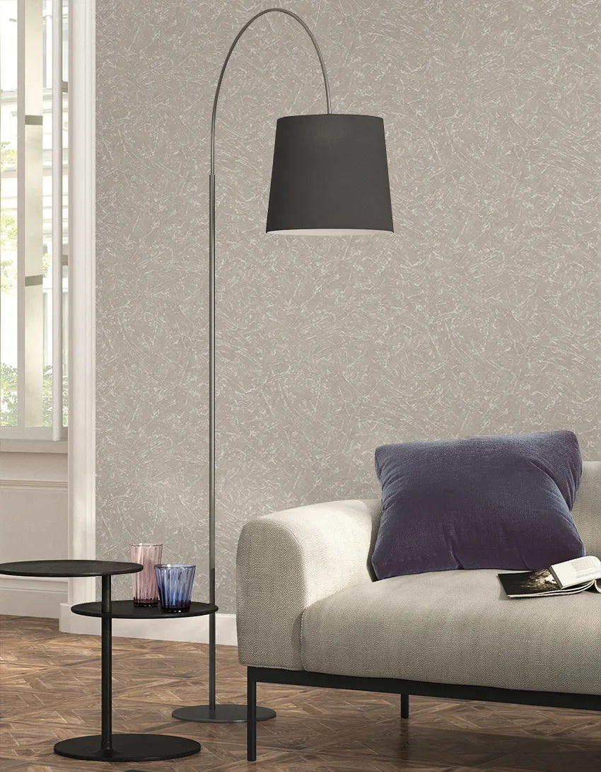 Marburg Loft Superior 34154 Beige wallpaper with a subtle abstract streak pattern behind a modern living room setup with an arched floor lamp, a light sofa, and a round side table with glasses.