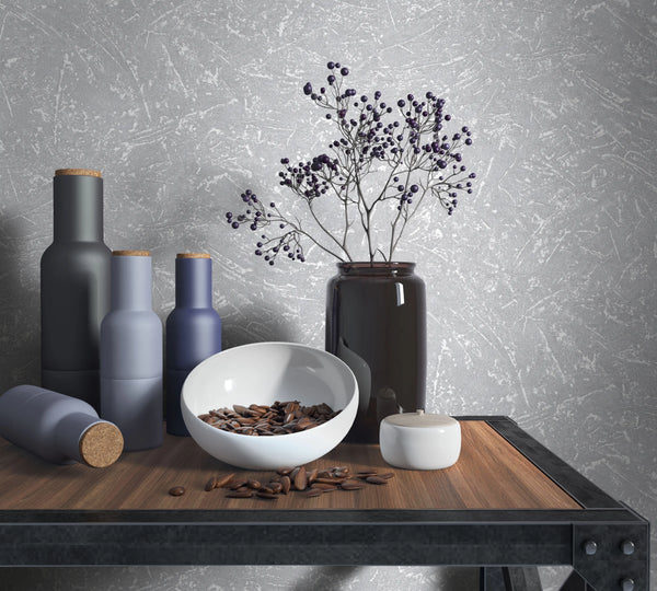 Marburg Loft Superior 34155 Gray textured wallpaper with an abstract plaster-like pattern behind a wood-and-metal table holding matte bottles with cork stoppers, a dark glass vase with berry stems, a white bowl with seeds, and a small lidded jar.