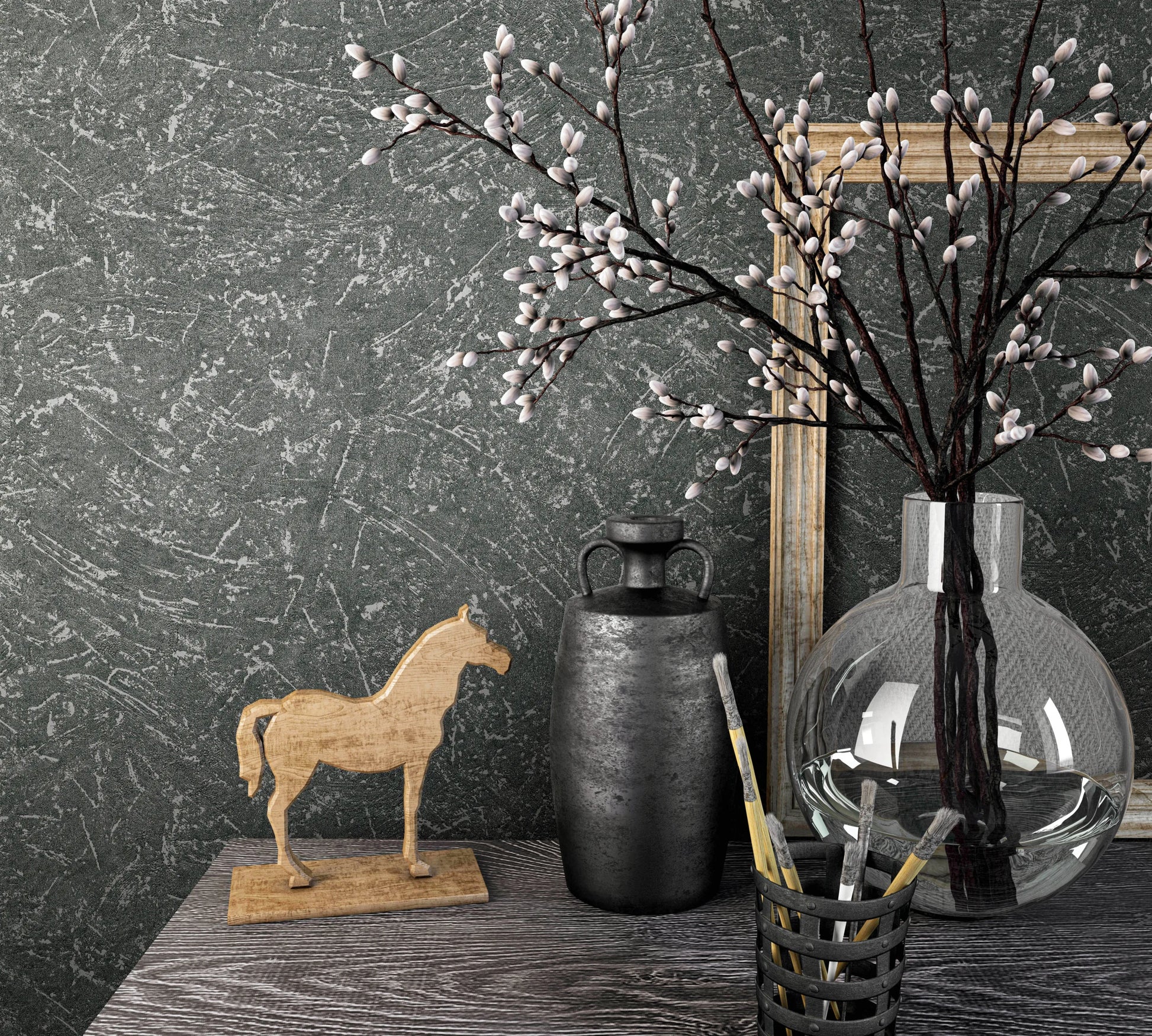 Marburg Loft Superior 34156 Dark gray textured wallpaper with a light gray scraped pattern behind a tabletop vignette featuring a round glass vase of budding branches, a dark metal jug, a wooden horse figurine, a cup of paintbrushes, and leaning wooden frames.