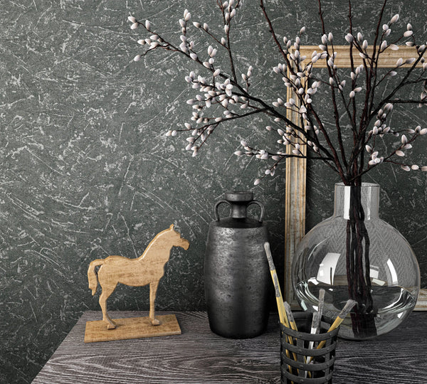 Marburg Loft Superior 34156 Dark gray textured wallpaper with a light gray scraped pattern behind a tabletop vignette featuring a round glass vase of budding branches, a dark metal jug, a wooden horse figurine, a cup of paintbrushes, and leaning wooden frames.