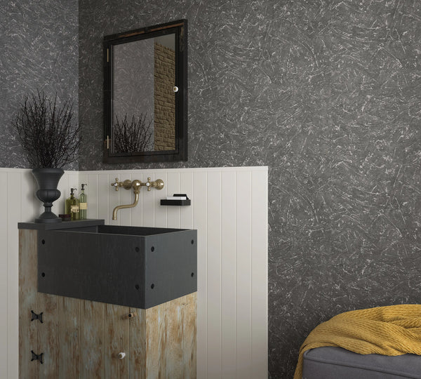 Marburg Loft Superior 34156 Dark anthracite wallpaper with an abstract scraped texture is shown above white vertical paneling in a bathroom, beside a vanity with a deep black sink, wall-mounted faucet, framed mirror, and a small ottoman with a blanket.