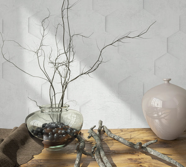 Marburg Loft Superior 34157 Light grey-white wallpaper with a subtle hexagonal relief pattern behind a wood tabletop styled with a glass vase of branches, scattered twigs, and a glossy ceramic vessel.