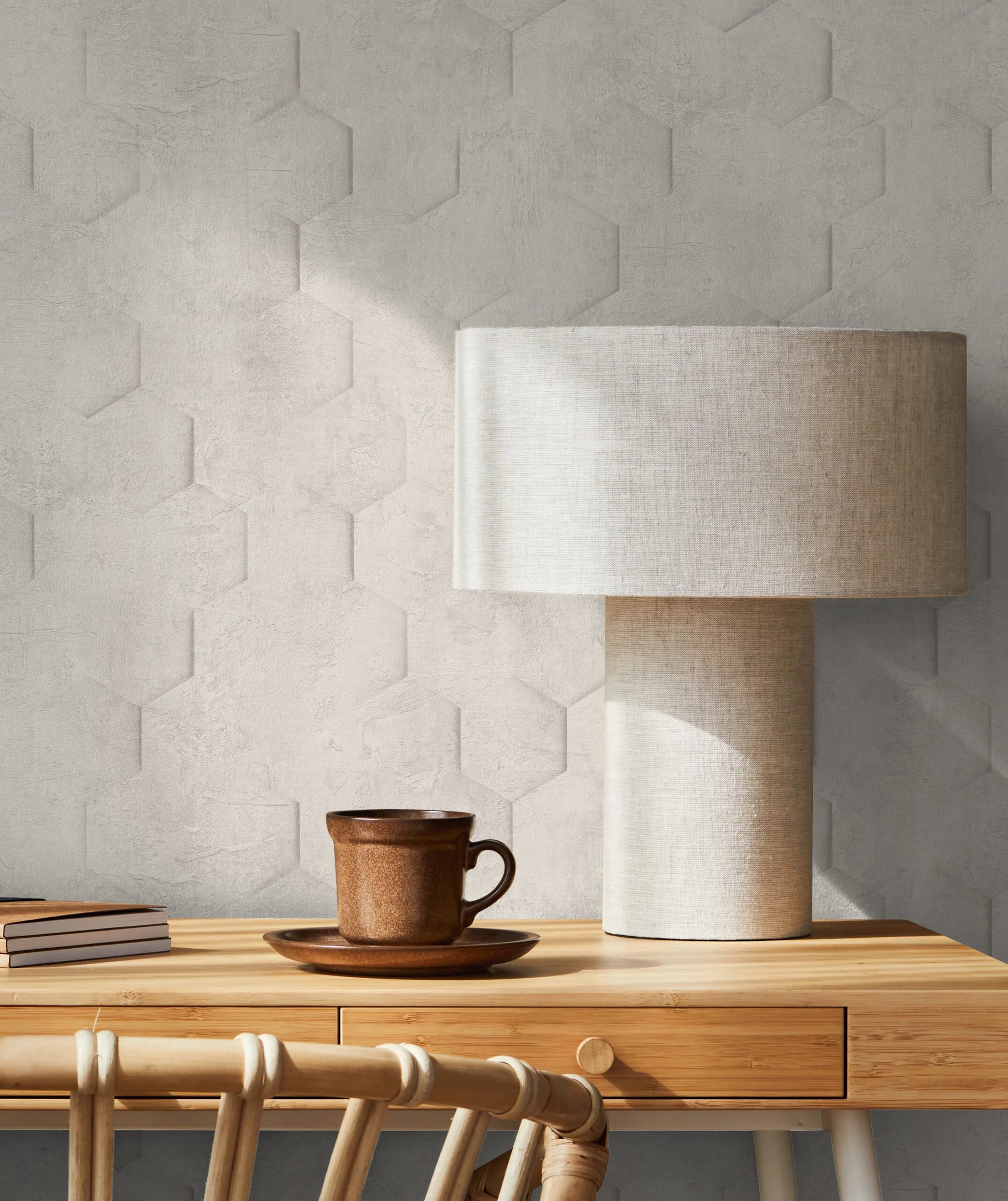 Marburg Loft Superior 34158 Tone-on-tone hexagonal embossed wallpaper behind a wooden desk with a fabric table lamp, a brown ceramic mug on a saucer, stacked notebooks, and a rattan chair.