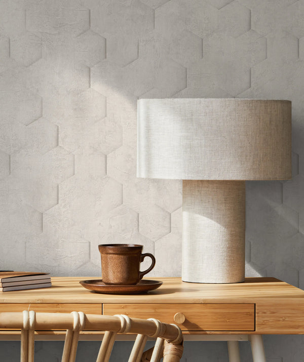 Marburg Loft Superior 34158 Tone-on-tone hexagonal embossed wallpaper behind a wooden desk with a fabric table lamp, a brown ceramic mug on a saucer, stacked notebooks, and a rattan chair.