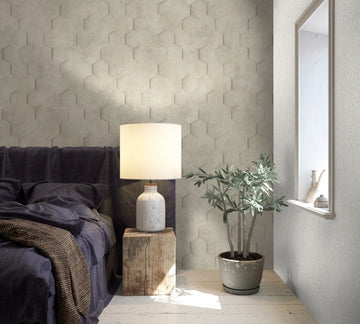 Marburg Loft Superior 34158 Light beige hexagon relief wallpaper behind a bed, with a stone-look table lamp on a wood block and a potted plant beside a window.