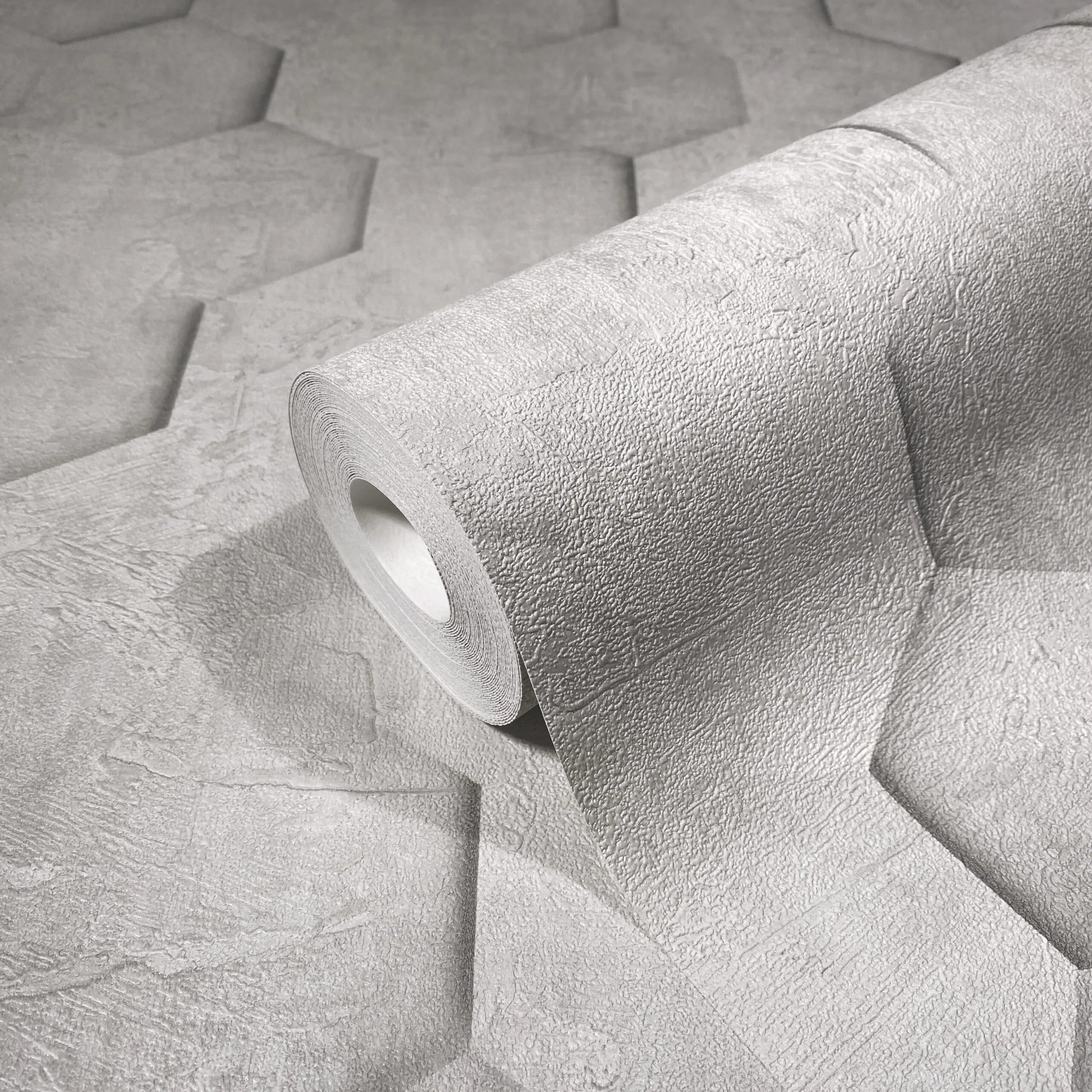 Marburg Loft Superior 34159 Close-up of a gray textured wallpaper roll resting on an unrolled sheet that shows a large hexagon panel pattern with a plaster-like finish.