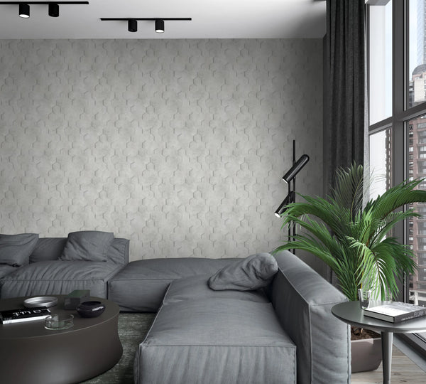 Marburg Loft Superior 34159 Light gray hexagon-pattern wallpaper with a subtle 3D effect covers the feature wall behind a gray sectional. A round coffee table, side table, black floor lamp, potted palm, tall windows with city view, curtains, and ceiling track lights are visible.