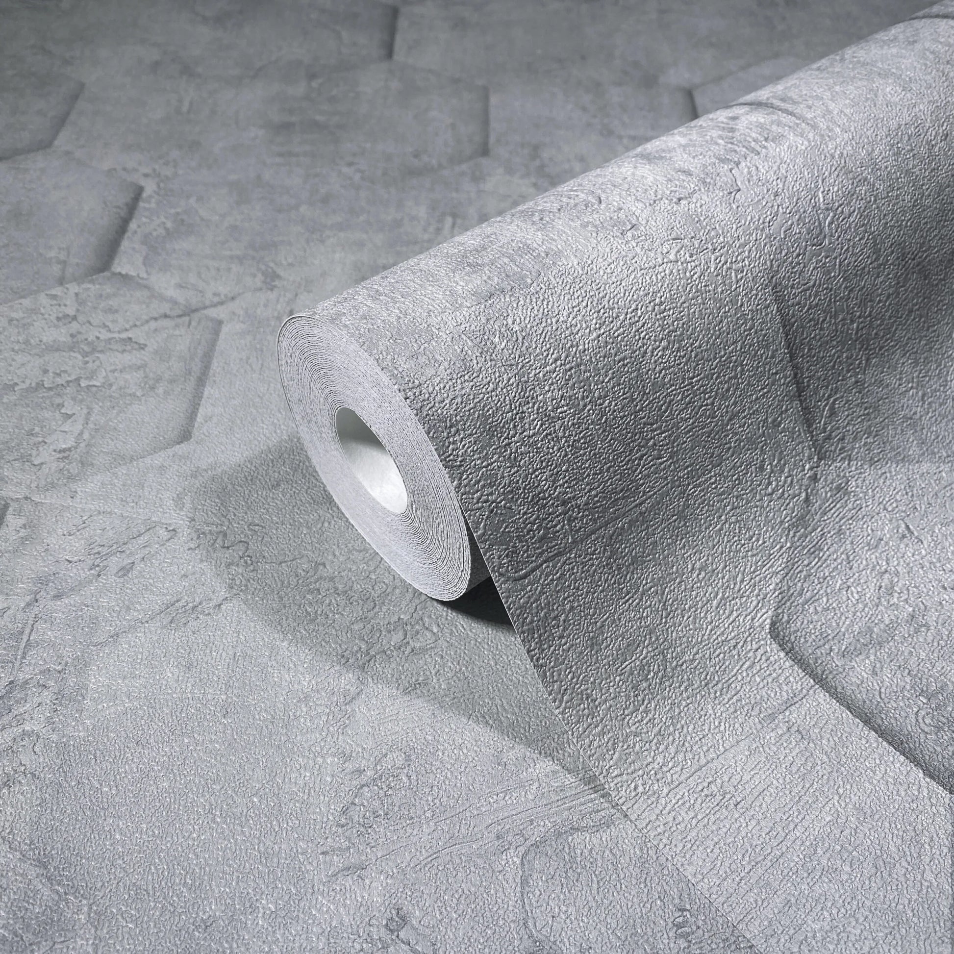 Marburg Loft Superior 34160 A gray textured wallpaper roll resting on the same pattern, showing an embossed stone-like surface with faint geometric shapes.