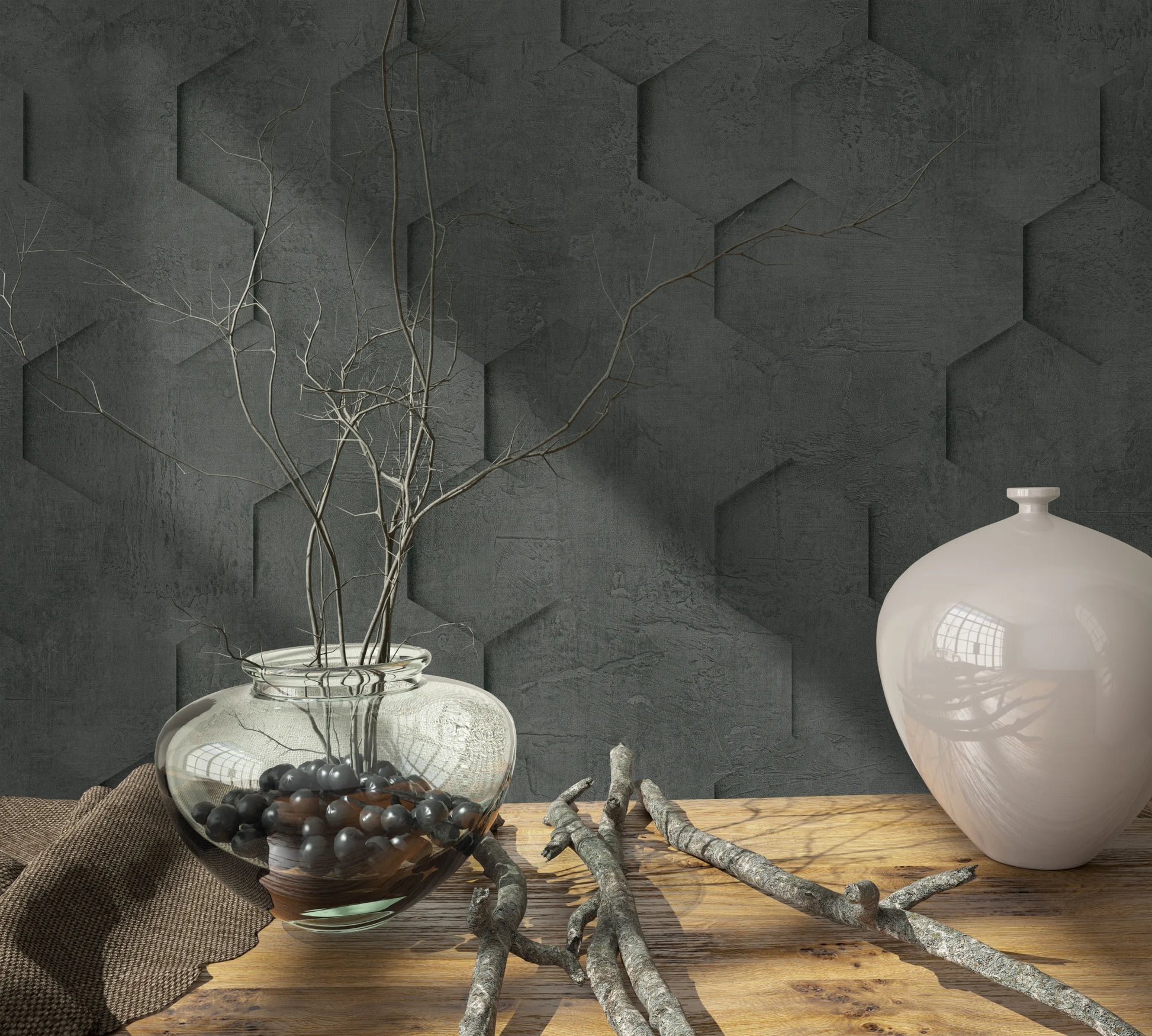 Marburg Loft Superior 34161 Dark charcoal geometric wallpaper with a concrete-like texture and a hexagon pattern that appears three-dimensional. In front, a wooden tabletop holds a clear glass bowl vase with branches and black stones, a glossy light ceramic vase, loose twigs, and a brown.