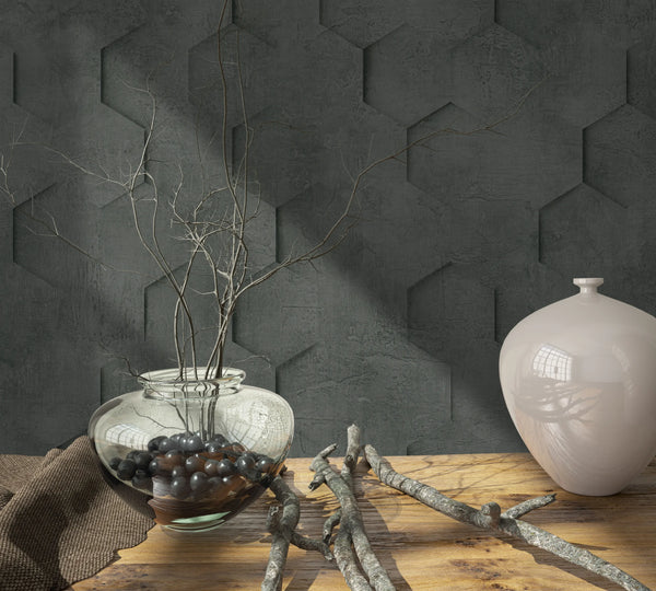 Marburg Loft Superior 34161 Dark charcoal geometric wallpaper with a concrete-like texture and a hexagon pattern that appears three-dimensional. In front, a wooden tabletop holds a clear glass bowl vase with branches and black stones, a glossy light ceramic vase, loose twigs, and a brown.