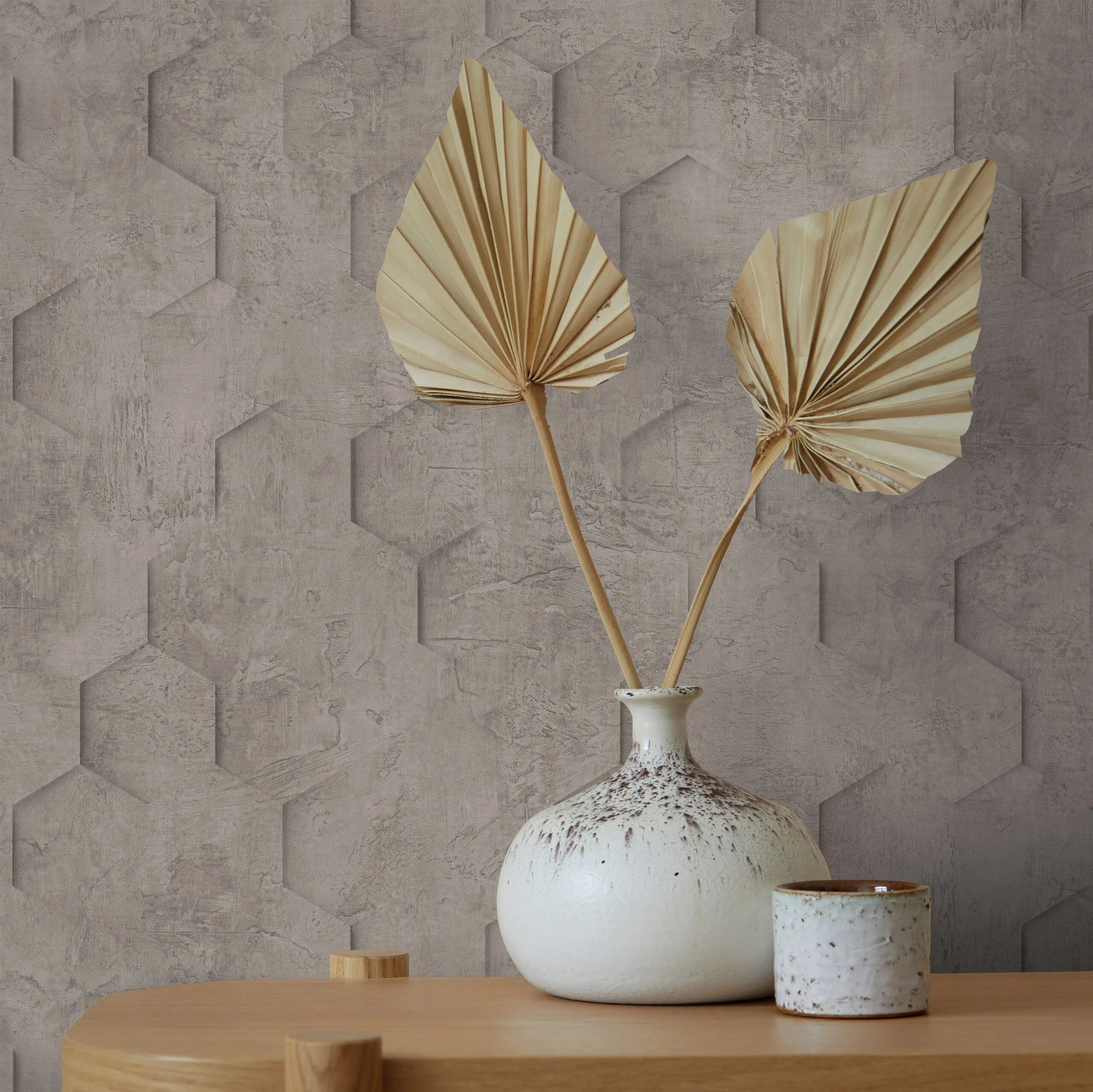 Marburg Loft Superior 34162 Taupe-brown geometric wallpaper with a concrete texture and subtle 3D hexagon pattern, shown behind a ceramic vase with dried leaves and a small cup on a wooden table.
