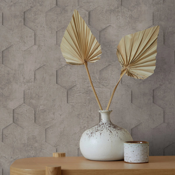 Marburg Loft Superior 34162 Taupe-brown geometric wallpaper with a concrete texture and subtle 3D hexagon pattern, shown behind a ceramic vase with dried leaves and a small cup on a wooden table.