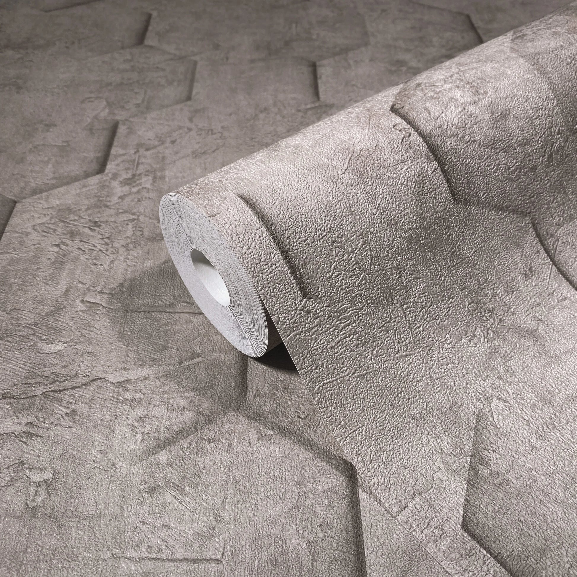Marburg Loft Superior 34162 A wallpaper roll resting on matching unrolled wallpaper with an embossed geometric pattern and stone-like texture in a neutral tone.