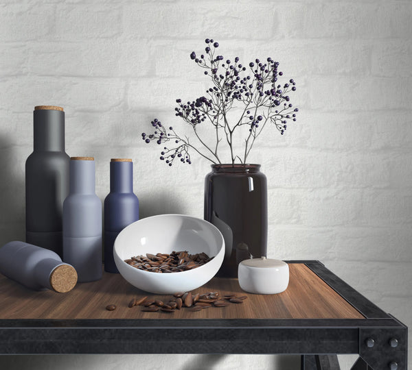 Marburg Loft Superior 34165 White brick-effect wallpaper behind a wood-and-metal table styled with matte bottles with cork stoppers, a dark glass vase holding berry stems, a white bowl with seeds, and a small lidded jar.