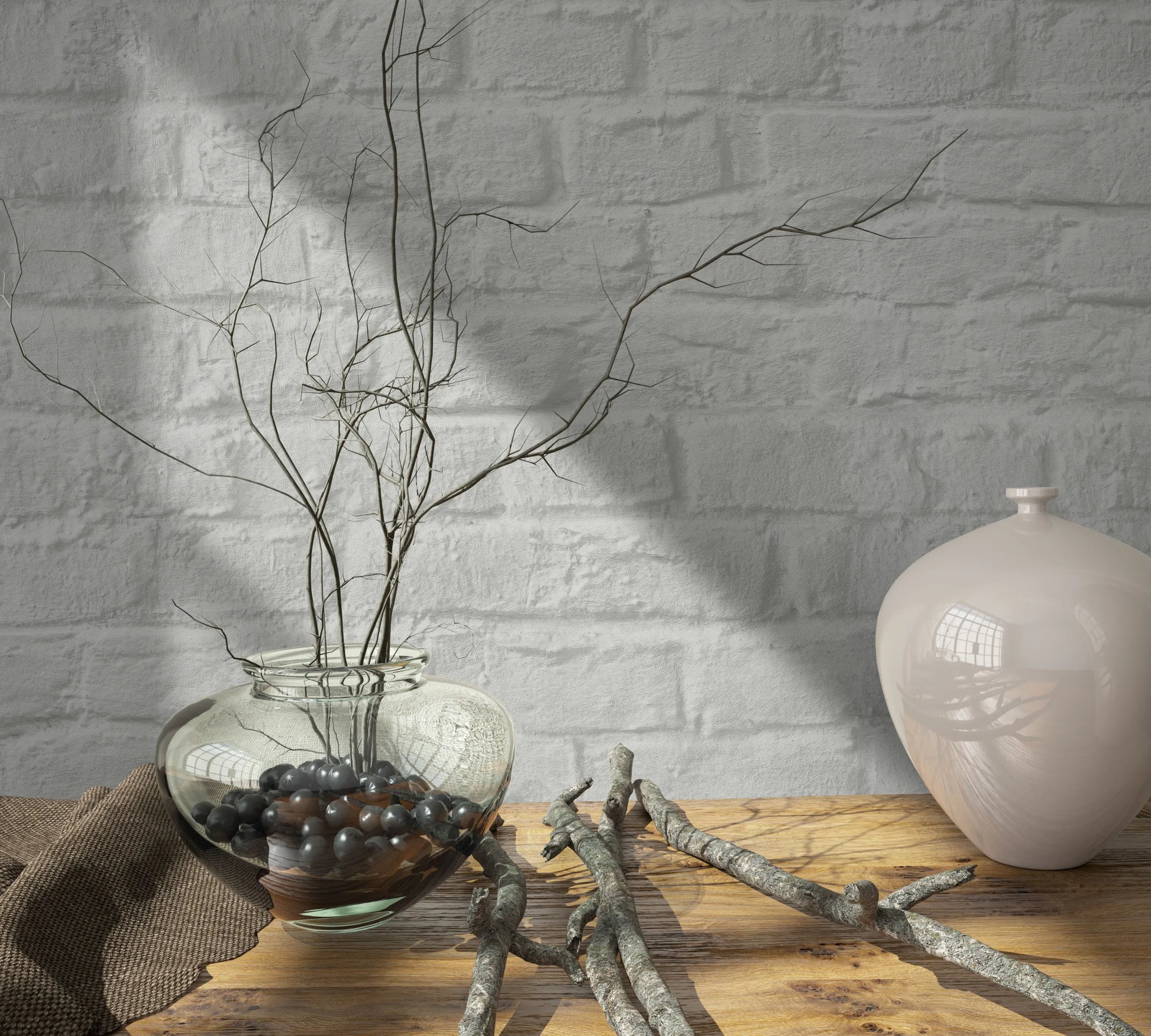 Marburg Loft Superior 34166 Light gray faux brick wallpaper behind a wooden tabletop styled with a clear glass vase holding twigs and dark decorative stones, loose branches, a brown cloth, and a glossy ceramic vase.